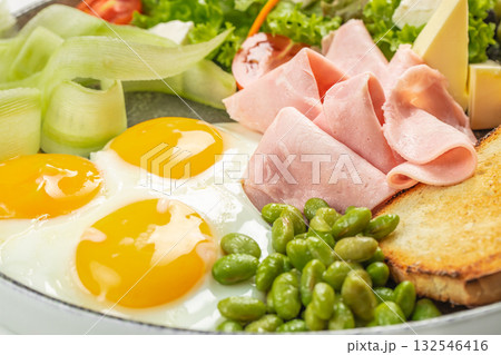 Close-up of a breakfast dish with fried eggs, sliced ham, toast, fresh vegetables, and edamame beans Close-up of a breakfast dish with fried eggs, sliced ham, toast, fresh vegetables, and edamame beans 132546416