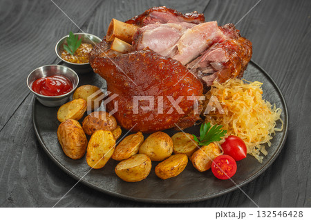 Juicy roasted pork knuckle served with golden baked potatoes, sauerkraut, cherry tomatoes, ketchup, and mustard on a black plate Juicy roasted pork knuckle served with golden baked potatoes, sauerkraut, cherry tomatoes, ketchup, and mustard on a black plate 132546428