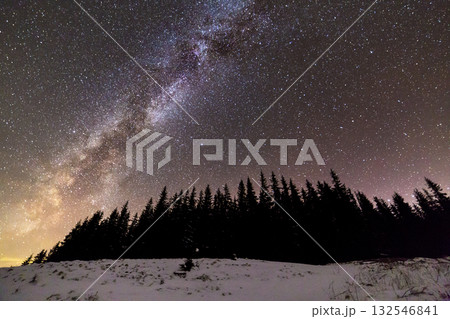 Winter mountains night landscape panorama. Milky Way bright constellation in dark blue starry sky over dark spruce pine trees forest, soft glow on horizon after sunset. Wide angle shot. 132546841