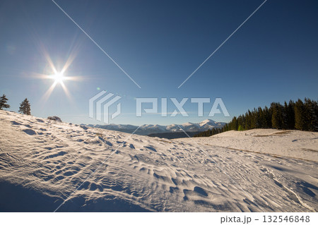 Winter landscape panorama with snowy landscape hills, distant white mountains, dark forest and clear blue sky with bright sun. 132546848