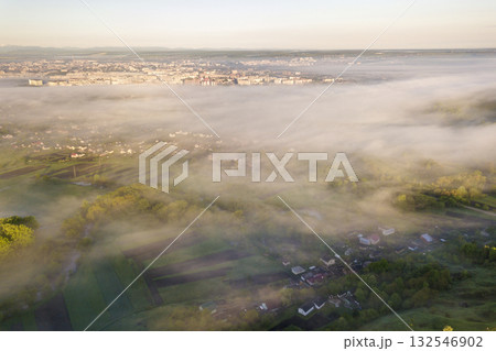 View from above of white fog on village house roofs among green trees under bright blue sky. Spring misty landscape panorama at dawn. View from above of white fog on village house roofs among green trees under bright blue sky. Spring misty landscape panorama at dawn. 132546902