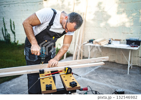 Mature male carpenter using electric drill on a wooden plank outdoors 132546982