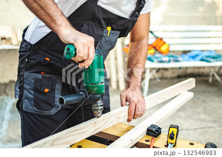 Unrecognizable male carpenter using electric drill on a wooden plank outdoors Unrecognizable male carpenter using electric drill on a wooden plank outdoors 132546983