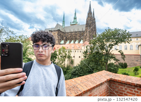 Prague, Czech Republic, August 8, 2023. A young man takes a selfie with his cell phone at the view of Prague Castle and St. Vitus Cathedral on the hill. A beautiful summer day. Travel destinations. Prague, Czech Republic, August 8, 2023. A young man takes a selfie with his cell phone at the view of Prague Castle and St. Vitus Cathedral on the hill. A beautiful summer day. Travel destinations. 132547420