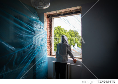 Quiet renovation contemplation, Hooded figure surveys ongoing window renovation activities from window overlooking leafy street 132548413