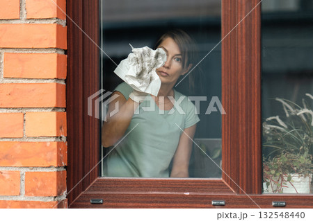 Lady wiping window with concentrated expression, Caucasian woman in casual attire carefully cleaning window in cozy home Lady wiping window with concentrated expression, Caucasian woman in casual attire carefully cleaning window in cozy home 132548440