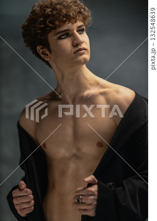 young man with curly hair and a black jacket stands in a studio looking upward with a confident expression. He displays his bare chest and emphasizes his striking features. young man with curly hair and a black jacket stands in a studio looking upward with a confident expression. He displays his bare chest and emphasizes his striking features. 132548639