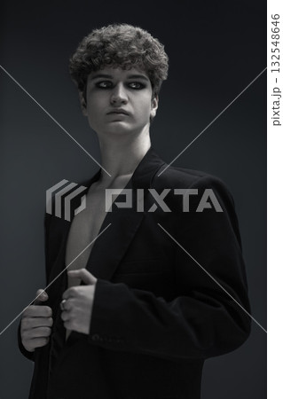 young man with curly hair stands confidently in a black jacket revealing his bare chest. The low lighting creates a dramatic atmosphere around him in the studio. 132548646