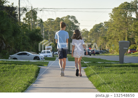 Rear view of two young teenage children, girl and boy, brother and sister walking together on rural street on bright sunny day. Vacation time concept 132548650