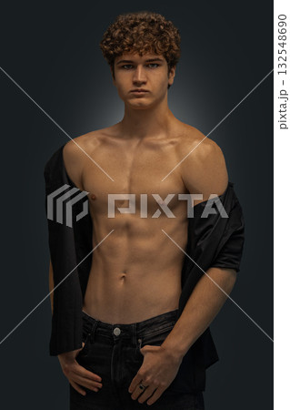 young man stands in a studio showcasing his curly hair and toned physique. He wears a black shirt casually draped over one shoulder while looking directly at the camera. young man stands in a studio showcasing his curly hair and toned physique. He wears a black shirt casually draped over one shoulder while looking directly at the camera. 132548690