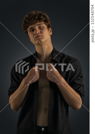 young man showcases a confident expression while adjusting his black shirt. The studio lighting highlights his features creating a striking contrast with the dark background. young man showcases a confident expression while adjusting his black shirt. The studio lighting highlights his features creating a striking contrast with the dark background. 132548704