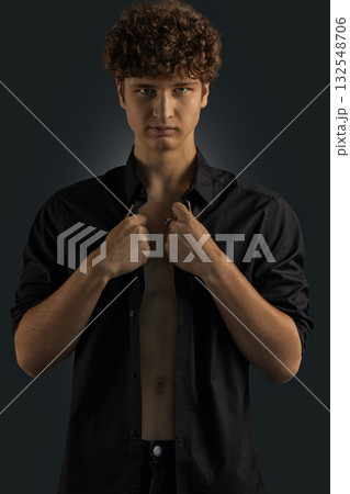 Curly haired young man stands in a black shirt giving a confident look. He is indoors against a dark backdrop with a focus on his expression and pose. Curly haired young man stands in a black shirt giving a confident look. He is indoors against a dark backdrop with a focus on his expression and pose. 132548706