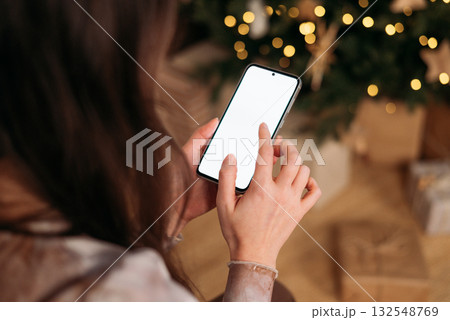 A woman's hands using a vertical smartphone with a blank white screen, blurred festive Christmas tree and gifts A woman's hands using a vertical smartphone with a blank white screen, blurred festive Christmas tree and gifts 132548769