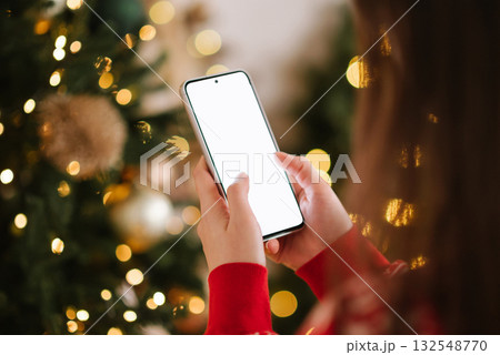 A close-up of a person's hands in a red sweater holding a vertical smartphone with a bright white blank screen against a background of sparkling Christmas bokeh. A close-up of a person's hands in a red sweater holding a vertical smartphone with a bright white blank screen against a background of sparkling Christmas bokeh. 132548770