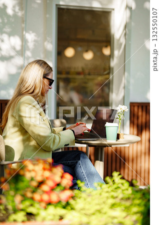 Woman working on laptop at outdoor cafe surrounded by plants and flowers in warm sunlight Woman working on laptop at outdoor cafe surrounded by plants and flowers in warm sunlight 132550107