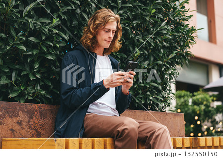 Young man checks his phone while sitting on a bench in an urban setting during daytime Young man checks his phone while sitting on a bench in an urban setting during daytime 132550150