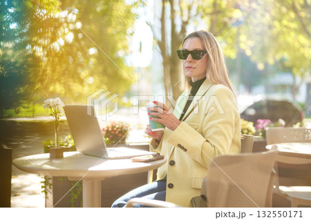 Young woman working on laptop outside while enjoying coffee at a sunny cafe Young woman working on laptop outside while enjoying coffee at a sunny cafe 132550171