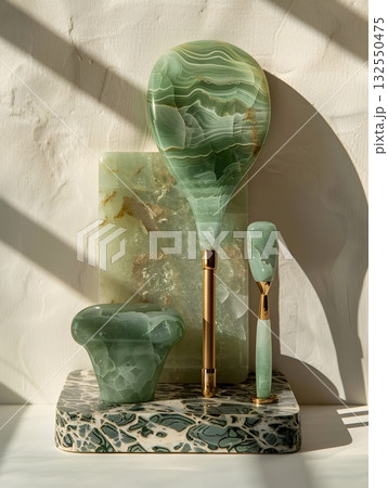Minimal beauty setup featuring a polished jade gua-sha board, facial roller on a gold stand, oval massage stone and jade wand arranged on a textured stone tray. Clean light and marble veins emphasize  132550475