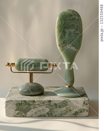 Minimal beauty setup featuring a polished jade gua-sha board, facial roller on a gold stand, oval massage stone and jade wand arranged on a textured stone tray. Clean light and marble veins emphasize  132550488