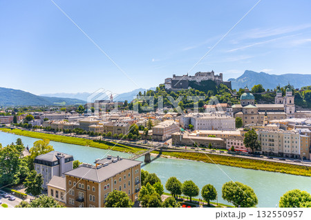 This stunning view shows Salzburg, Austria, featuring the iconic castle atop the hill. The blue sky reflects over the river, creating a serene atmosphere. 132550957