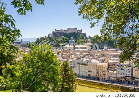 Morning light brightens the historic streets of Salzburg. The impressive fortress looms above the colorful buildings by the Danube, surrounded by lush green trees and blue skies. 132550959
