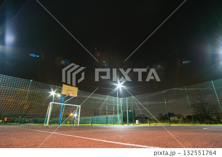 Outdoors mini football and basketball court with ball gate and basket surrounded with high protective fence brightly illuminated with spotlight lamps at night. Outdoors mini football and basketball court with ball gate and basket surrounded with high protective fence brightly illuminated with spotlight lamps at night. 132551764