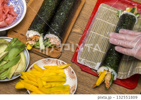 Making sushi and rolls at home. Plates with ingredients for traditional Japanese food and sushi rolls on wooden board on kitchen table, view from above. Making sushi and rolls at home. Plates with ingredients for traditional Japanese food and sushi rolls on wooden board on kitchen table, view from above. 132551809