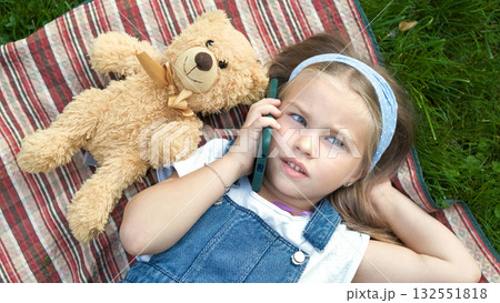 Little cute child girl laying on a blanket on green lawn in summer outdoors with her teddy bear toy talking on mobile phone. Little cute child girl laying on a blanket on green lawn in summer outdoors with her teddy bear toy talking on mobile phone. 132551818