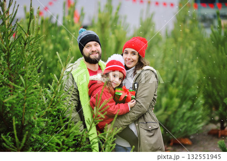 parents hold their little daughter in their arms with a Christmas present in their hands and smile against the backdrop of many Christmas trees and snow. parents hold their little daughter in their arms with a Christmas present in their hands and smile against the backdrop of many Christmas trees and snow. 132551945
