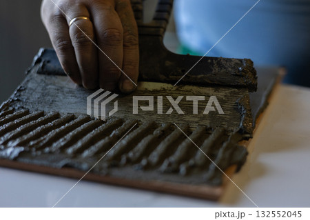 A person's hand applies mortar with a notched trowel to the back of a tile. A close-up of textured adhesive and the skillful work of tiling during a home renovation or flooring installation. 132552045