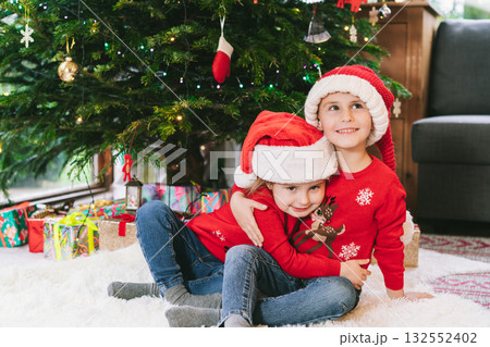 Two smiling kids in festive sweaters and Santa hats sitting under Christmas tree at home during holiday. Happy sister cuddling brother, feeling excited, having fun, childhood miracle time concept Two smiling kids in festive sweaters and Santa hats sitting under Christmas tree at home during holiday. Happy sister cuddling brother, feeling excited, having fun, childhood miracle time concept 132552402