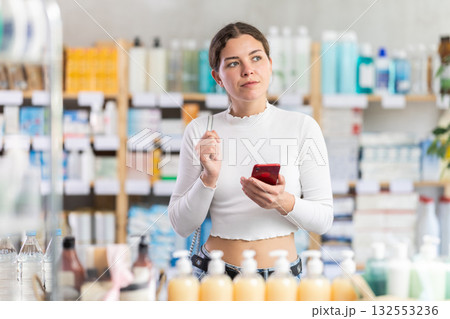 Female shopper searches for necessary goods on the shelves of a supermarket, checking the list on smartphone 132553236