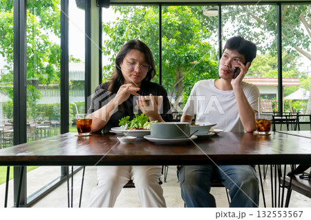 Two Thai LGBTQ friends enjoying lunch together one checking photos while the other on a phone call in a cozy cafe showing friendship relaxation and real daily life moments 132553567
