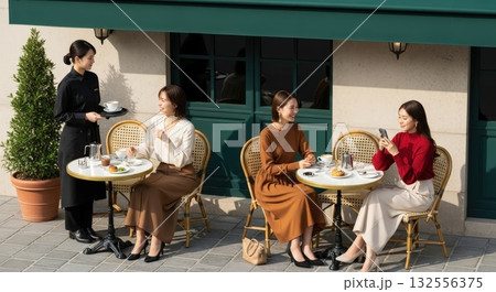 歩道目線で撮影した深緑オーニング街角カフェテラス、会話やスマホでくつろぐ女性 132556375