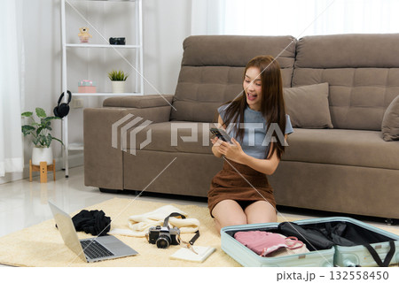 Young woman happy home activity preparing travel, packing luggage, using smartphone for booking, laptop for planning, vacation anticipation 132558450