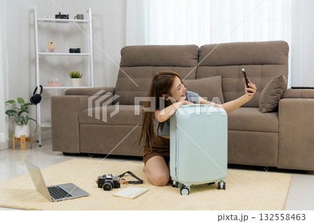 Young happy woman home activity shows people planning trip, posing for smartphone selfie, leaning on light blue luggage, laptop and vintage camera ready, excited for adventure Young happy woman home activity shows people planning trip, posing for smartphone selfie, leaning on light blue luggage, laptop and vintage camera ready, excited for adventure 132558463
