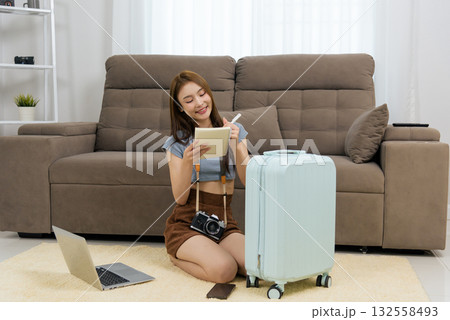 Young woman at home, happy people planning travel activity, writing ideas in notebook using pen, vintage camera, laptop, phone and suitcase nearby, excited for future journey Young woman at home, happy people planning travel activity, writing ideas in notebook using pen, vintage camera, laptop, phone and suitcase nearby, excited for future journey 132558493