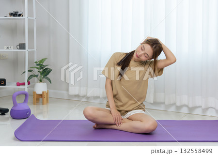 Young woman at home enjoys healthy activity, stretching neck on purple yoga mat, feeling happy, practicing mindful exercise for relaxation and body wellness among fitness equipment Young woman at home enjoys healthy activity, stretching neck on purple yoga mat, feeling happy, practicing mindful exercise for relaxation and body wellness among fitness equipment 132558499