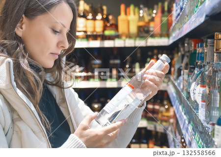 A woman with long hair examines the label on a vodka bottle while standing in the alcoholic beverages section of a supermarket. She appears focused on her choice. A woman with long hair examines the label on a vodka bottle while standing in the alcoholic beverages section of a supermarket. She appears focused on her choice. 132558566