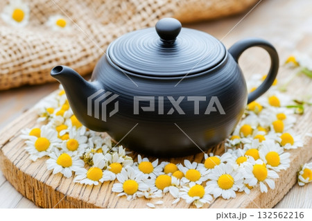 Rustic black teapot surrounded by dried chamomile flowers on weathered wooden surface creating a peaceful, aromatic atmosphere Rustic black teapot surrounded by dried chamomile flowers on weathered wooden surface creating a peaceful, aromatic atmosphere 132562216