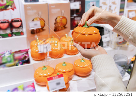 A woman chooses an orange ceramic pumpkin in a local market while shopping for Halloween gifts. The festive atmosphere features various seasonal decorations and souvenirs. A woman chooses an orange ceramic pumpkin in a local market while shopping for Halloween gifts. The festive atmosphere features various seasonal decorations and souvenirs. 132565251