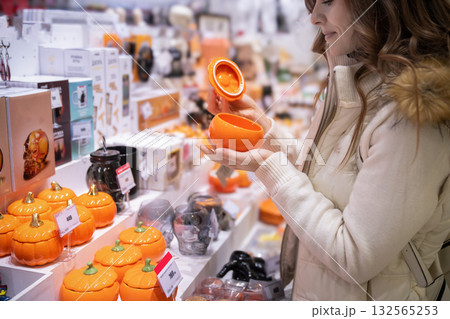 On the eve of Halloween, a woman browses a local market, selecting colorful ceramic pumpkins and unique jars for home decor. She enjoys the festive shopping atmosphere. On the eve of Halloween, a woman browses a local market, selecting colorful ceramic pumpkins and unique jars for home decor. She enjoys the festive shopping atmosphere. 132565253
