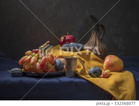 Still Life With Grapes, Apples, Plums, a Metal Cup, and Ornate Pitcher on Folded Fabric 132568077