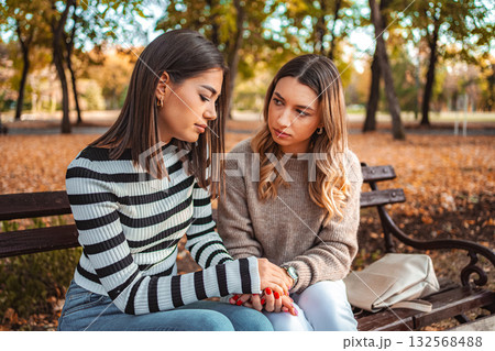 Two girls sit closely on a park bench surrounded by vibrant autumn leaves Two girls sit closely on a park bench surrounded by vibrant autumn leaves 132568488