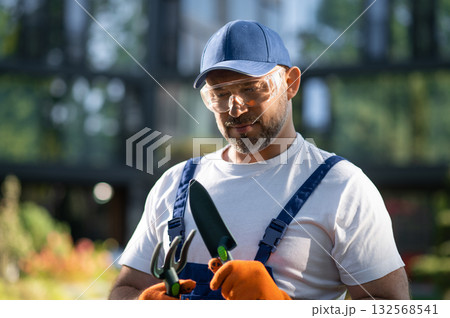 Professional gardener in uniform holding tools while standing in green garden Professional gardener in uniform holding tools while standing in green garden 132568541