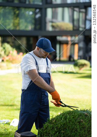 Man worker trimming greenery in landscaped yard of modern architecture complex Man worker trimming greenery in landscaped yard of modern architecture complex 132568548