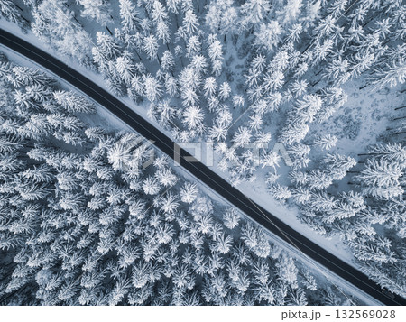 A winding road cuts through a winter landscape, surrounded by tall, snow-laden trees. The pristine white scenery creates a serene atmosphere, typical of a cold season. 132569028
