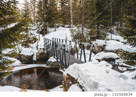 A picturesque winter scene showcases a creek in the Czech Republic known as Jizerka. Snow blankets the ground, and a bridge spans the cold waters, creating a tranquil atmosphere. 132569034