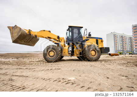 A large loader is working on a sandy area next to residential buildings. The overcast sky adds a muted backdrop to the scene A large loader is working on a sandy area next to residential buildings. The overcast sky adds a muted backdrop to the scene 132571164
