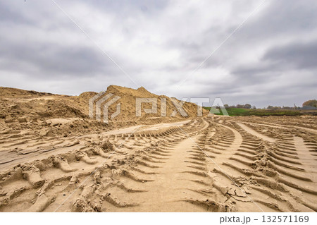 Deep tracks from heavy machinery cut through the sandy ground on a cloudy day. A large pile of sand rises in the background, hinting at ongoing construction 132571169